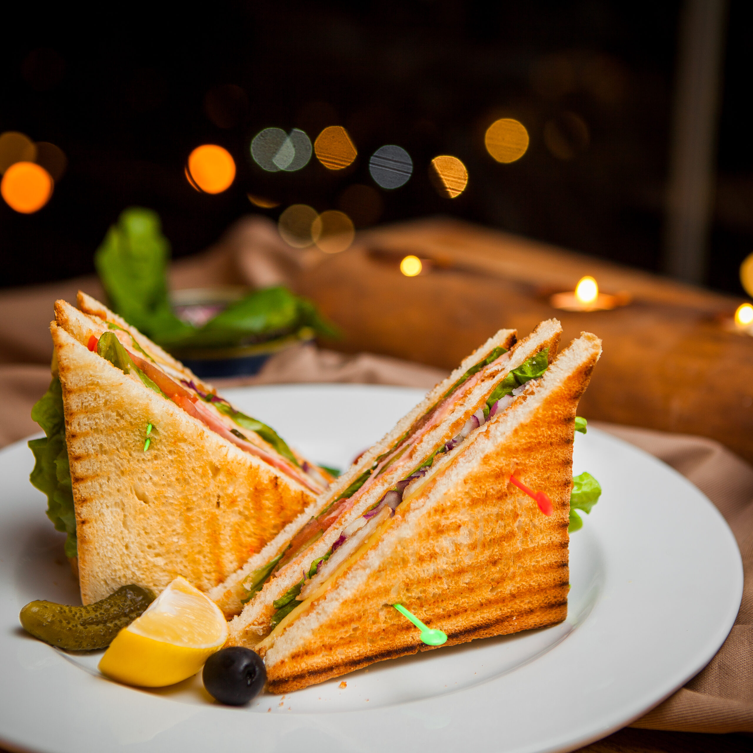 Side view club sandwich with salted cucumbers and lemon and olives in round white plate on wooden background
